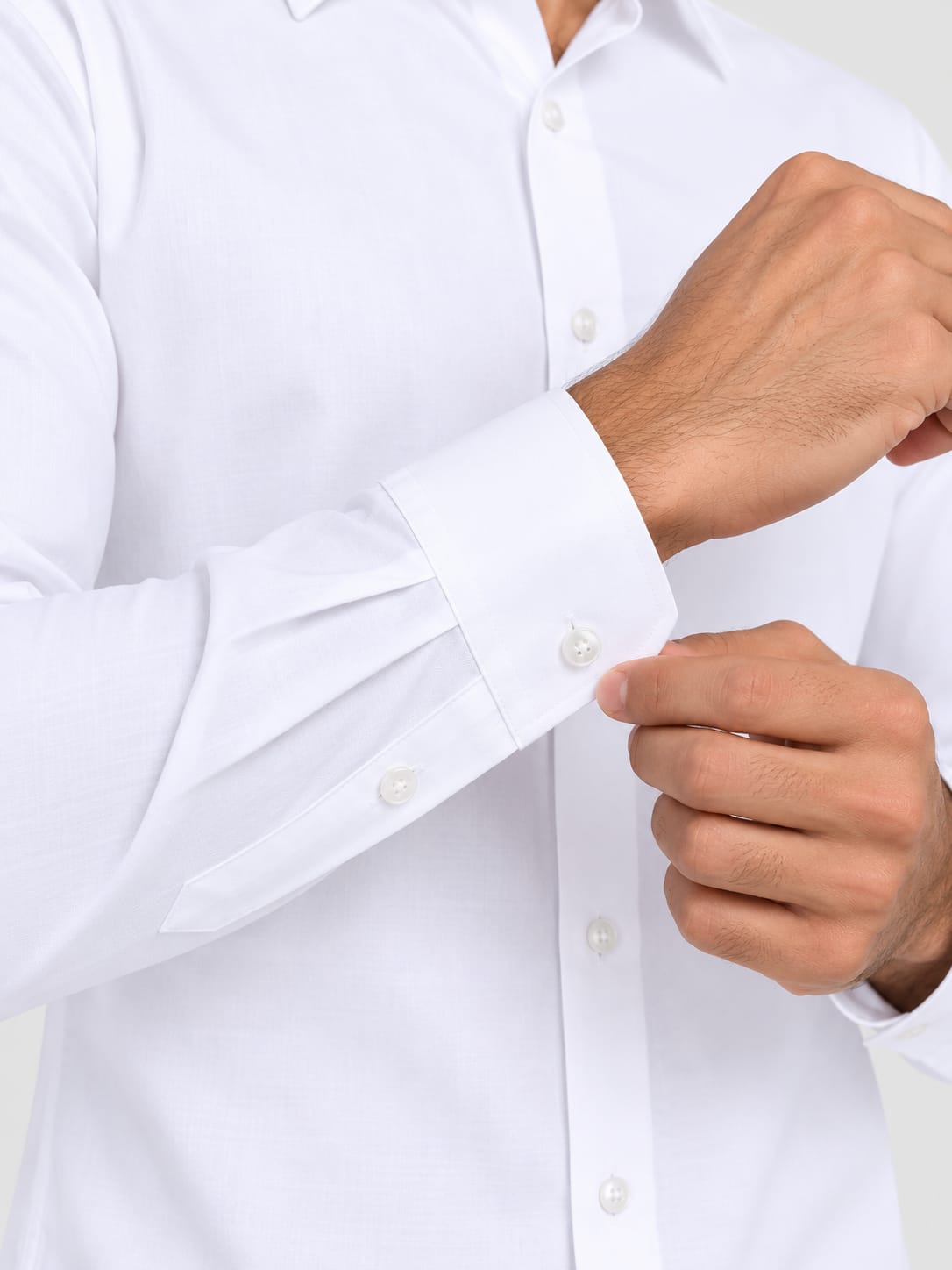 Close-up of a man's hands buttoning a white long-sleeve dress shirt. The image highlights the clean white fabric, the buttoned cuffs, and the front placket of the shirt against a light gray background.