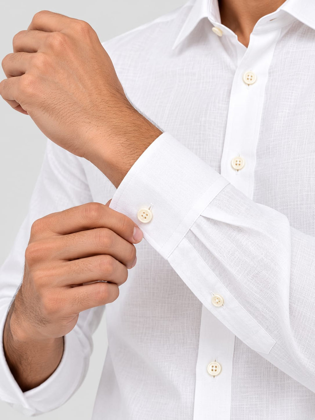 A close-up of a man's hands adjusting the cuff of a white linen button-down shirt. The shirt features a textured weave and cream-colored buttons, shown against a clean, light gray background.