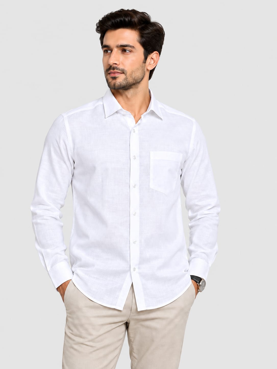 A man wearing a white long-sleeve button-down shirt with a chest pocket, paired with beige trousers, standing against a light gray studio background.
